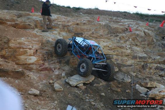 Buggy with blue roll cage climbing rocky hill. Person watches from above