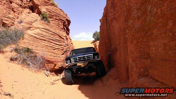 Black Ford Bronco navigating a narrow sandstone canyon with large tires and winch. The vehicle is angled, with its front end visible