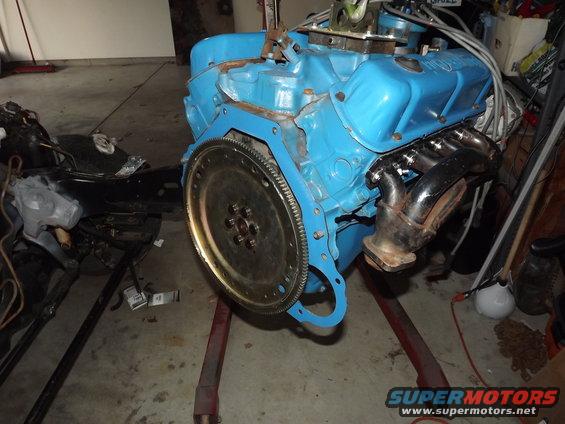 Blue engine block with black exhaust manifold and flywheel, mounted on an engine stand