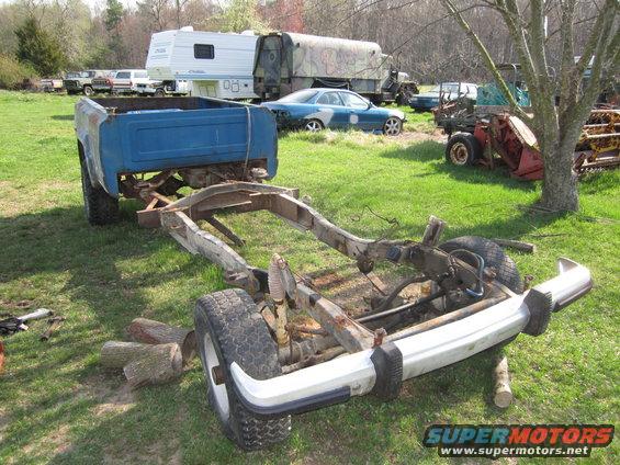 Truck chassis and frame with blue bed in background, other vehicles and RV in yard