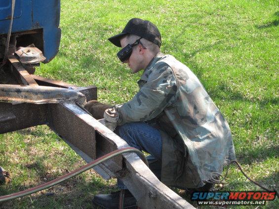 guy in camo jacket and cap welding on a rusty vehicle frame outdoors