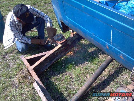 Man wearing a cap and plaid shirt welds a rusty metal support under a blue vehicle body