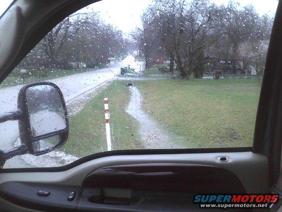 View from inside a vehicle looking out the window at a wet road and yard. Raindrops on the window. Side mirror visible