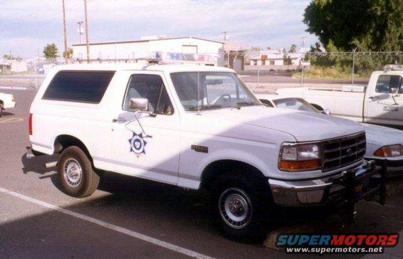 White 1994 Ford Bronco with police lights and a badge on the door, parked next to a pickup truck