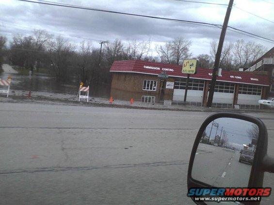 View of an auto repair shop with 'EMISSION CONTROL' and 'AUTO REPAIR' signs, next to a flooded area. A side mirror shows a car on the road