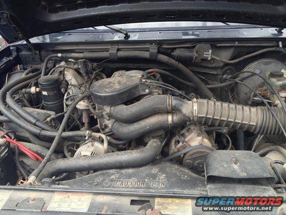 Engine bay with V8 engine, hoses, and fan shroud visible. Text on shroud reads 'CAUTION FAN'