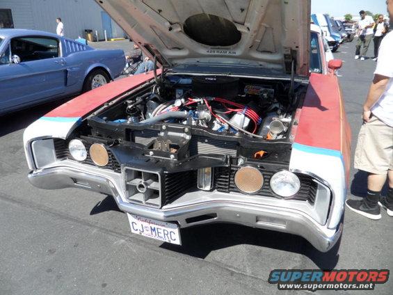 1970s Ford car front view, hood open showing engine. Another car is parked to the left