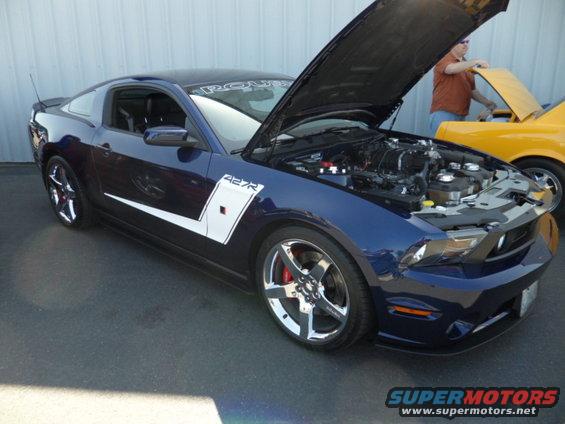 Blue Ford Mustang with hood open, showing engine. White stripe on side says 427R. Yellow car behind it
