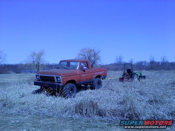 Red pickup truck with lifted suspension and off-road tires, parked in dry grass, with a green tractor in the background