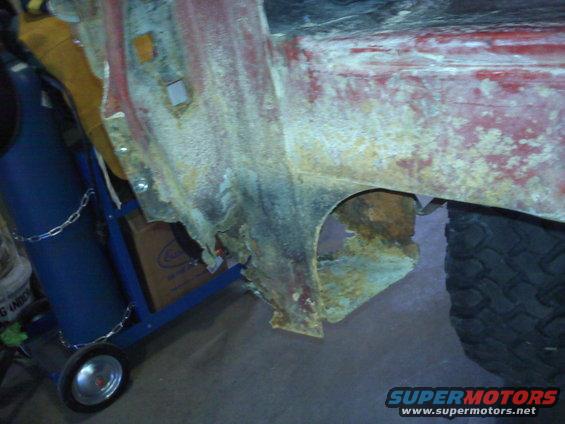 Heavily rusted red vehicle quarter panel with visible wheel well damage and a tire