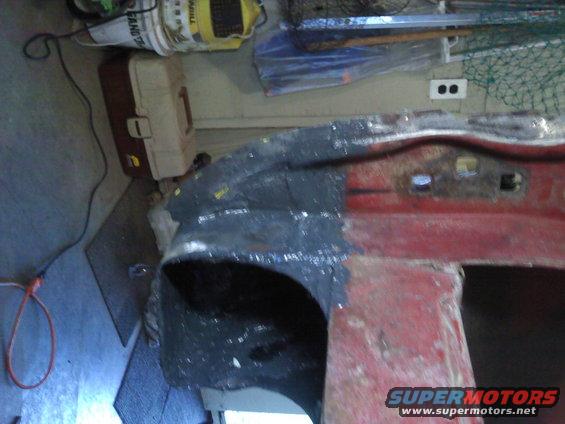Close-up of a red vehicle body panel being worked on, with gray primer applied in some areas. Tools and storage bins are visible in the background