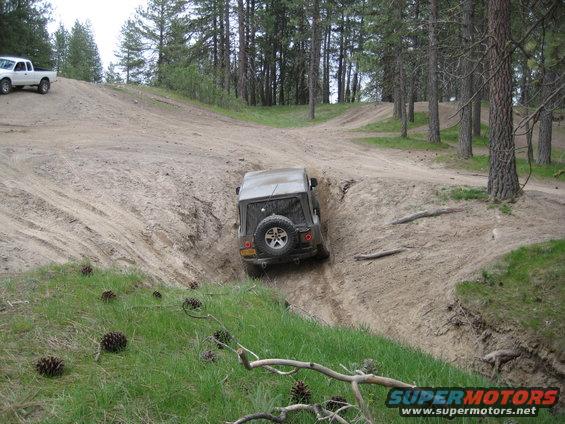 2005 Jeep Wrangler driving down a steep dirt embankment, white pickup truck on hill above