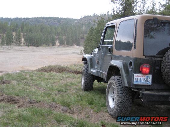 Jeep Wrangler parked on grassy hill overlooking a dirt area with trees in background. License plate reads ALT0508