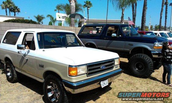 Two Ford Broncos parked outdoors, one white and tan, the other dark blue and gray with large tires
