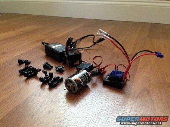 RC car motor, electronic speed controller, battery charger, and various black plastic suspension parts on a wooden floor