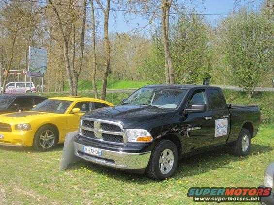 Black Dodge Ram 1500 pickup truck parked on grass, front 3/4 view. Yellow car to the left