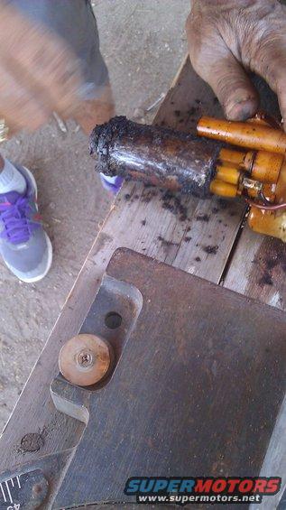 Dirty fuel pump assembly with orange connectors and a blackened cylindrical filter. A person's hands and feet are visible