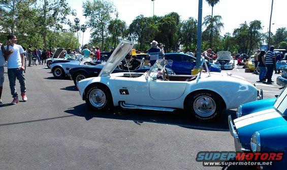 White convertible sports car with hood open, parked in a lot with other cars and people