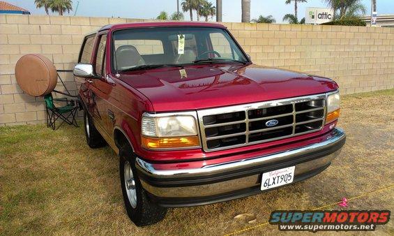 Red 1994 Ford Bronco parked on grass, front 3/4 view, chrome grille, tan spare tire cover on stand