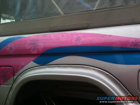 Side panel of a vehicle with pink and blue wavy graphics and floral patterns. Riveted fender flare visible