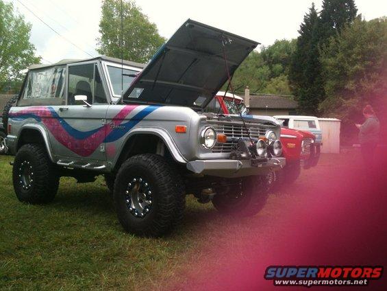 Silver Ford Bronco with pink and blue stripe graphics, hood open, lifted suspension, off-road tires