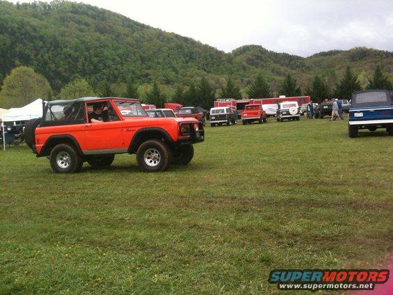 Orange first-gen Ford Bronco parked in a grassy field with other vehicles and tents in the background