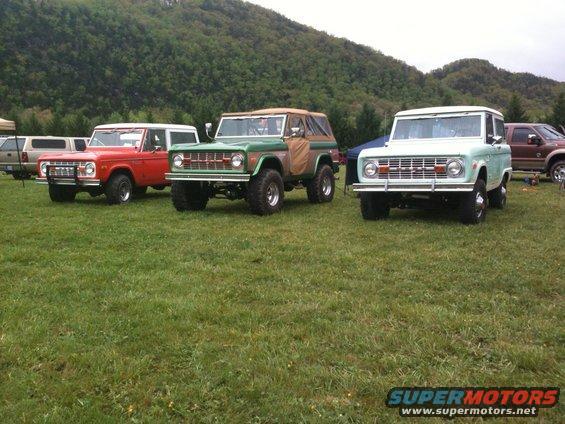 Three classic Ford Broncos parked on grass. The red Bronco appears to be from the early 1970s
