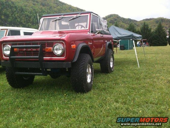 Red first-generation Ford Bronco parked on grass, another vehicle partially visible to the left