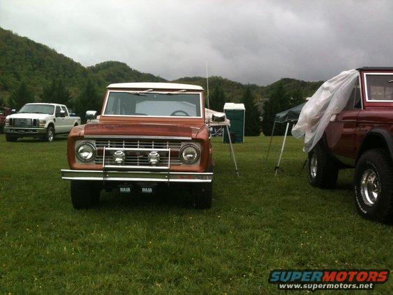 Front view of a brown Ford Bronco with a white top. Another Bronco is partially covered on the right. A white Ford pickup is in the background
