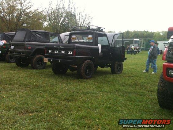 Rear view of a black Ford Bronco pickup truck with a soft top, parked on grass next to other vehicles