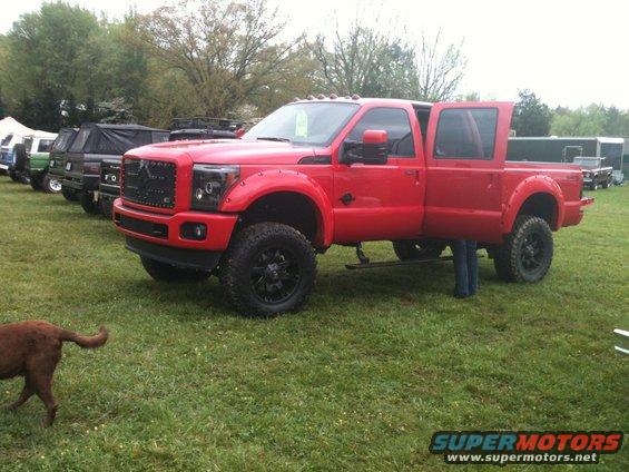 Red crew cab pickup truck with doors open, parked on grass with other vehicles in background. Dog in foreground