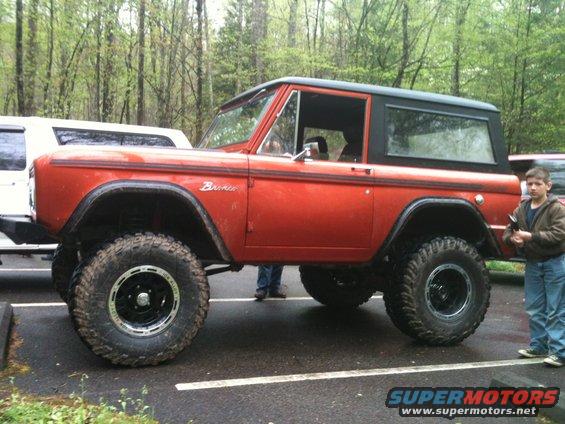 Orange Ford Bronco with black top, lifted suspension, and large tires. A boy stands to the right