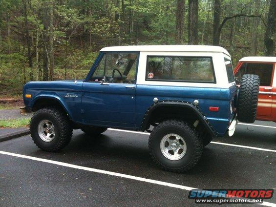 Blue 1978 Ford Bronco with white top, lifted suspension, and spare tire on rear. Another orange vehicle is parked next to it