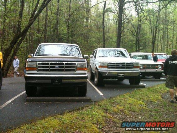 Two Ford Broncos parked side-by-side in a wooded area. The one on the left is dark, the one on the right is white