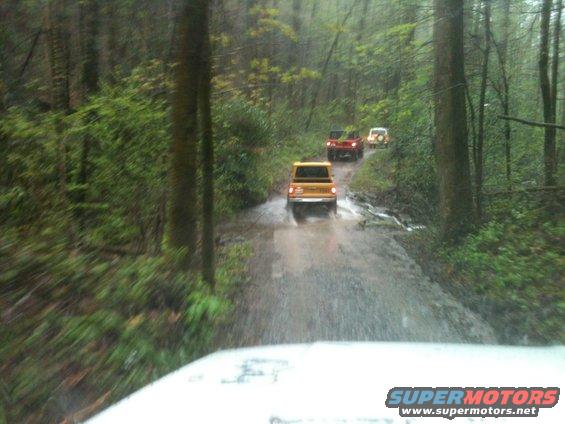 Yellow SUV leads red and white SUVs on a wet, muddy forest road