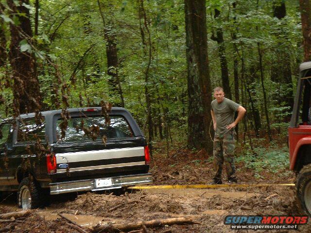 Ford Bronco stuck in mud with a man standing nearby and part of a red Jeep visible