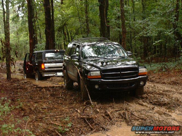 Black SUV in foreground, maroon SUV stuck in mud behind it, trees and mud everywhere