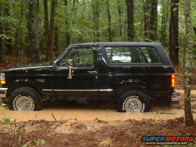 Black Ford Bronco stuck in mud, side view. The vehicle is partially submerged in a muddy trail surrounded by trees
