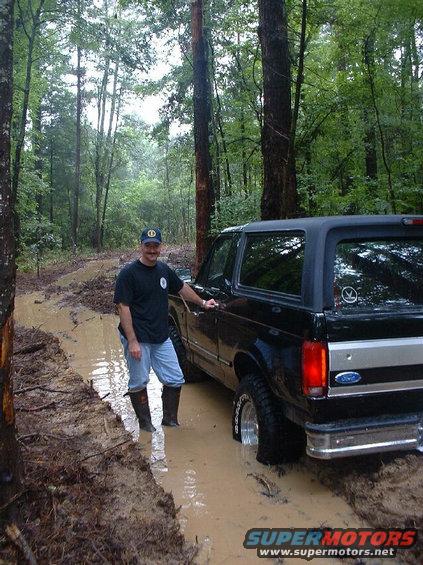 Man standing next to black Ford Bronco stuck in a muddy forest road. The vehicle is a 1992 model