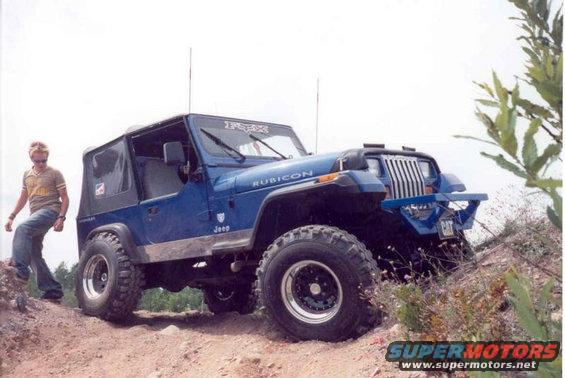 Blue Jeep Wrangler with large tires and a custom front bumper, parked on a dirt hill with a person standing nearby