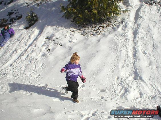 Children in winter clothing run down a snowy hill, one in the foreground, another in the background