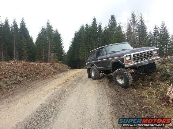 Black Ford Bronco with lifted suspension driving off-road on a dirt track through a forest. The grille and body style are visible