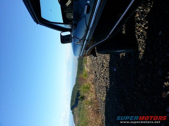 Side view of a dark SUV door and side mirror, parked on gravel with a wooded hillside and blue sky in background