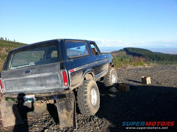 Dark blue 1978 Ford Bronco with lifted suspension and large tires, parked on a gravelly surface with a wooded hillside in the background