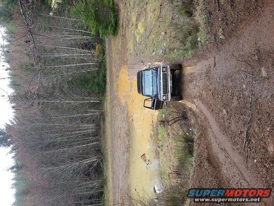 Dark blue 1978 Ford Bronco with driver's door open, driving through a muddy forest road