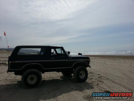 Black Ford Bronco parked on sand near ocean waves. Red flag on pole in background