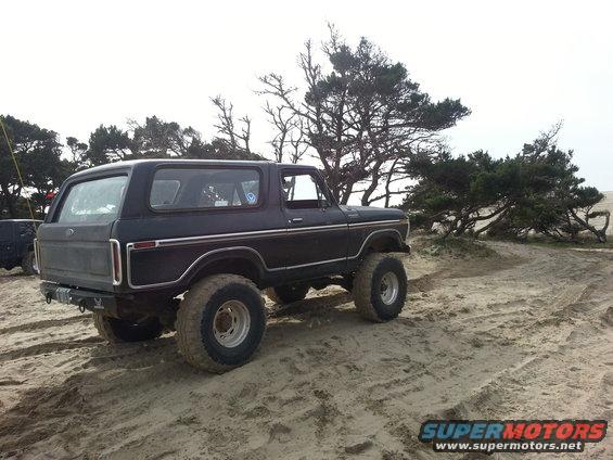 Dark SUV with off-road tires on sand, rear quarter view. Appears to be a 1978 Ford Bronco