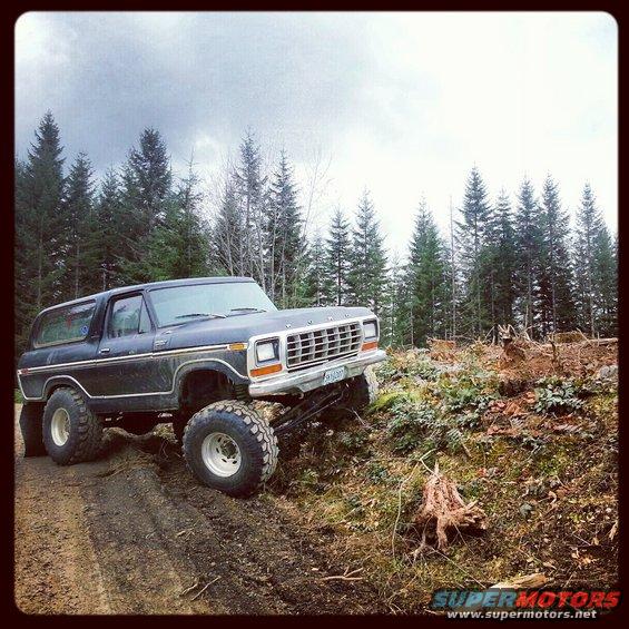 Dark blue Ford Bronco with lifted suspension and large tires on a muddy incline, surrounded by trees