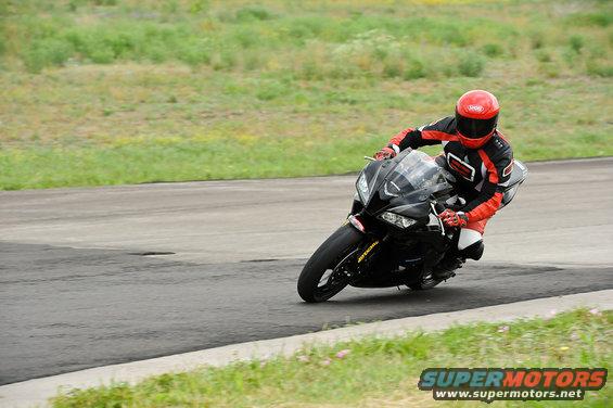 Rider in red and black gear leans into a turn on a black sportbike on a paved track with grassy shoulders