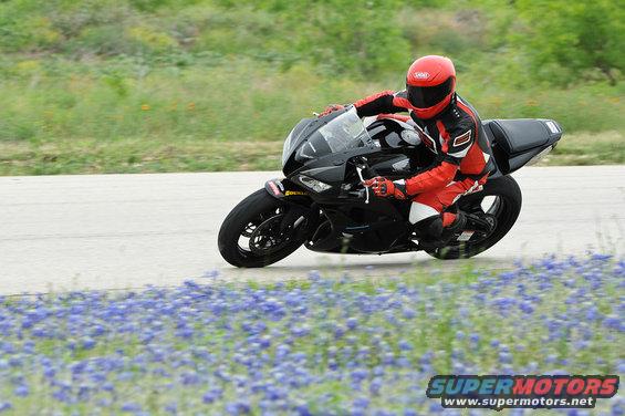 Black sport motorcycle with a rider in red gear on a paved track with bluebonnets